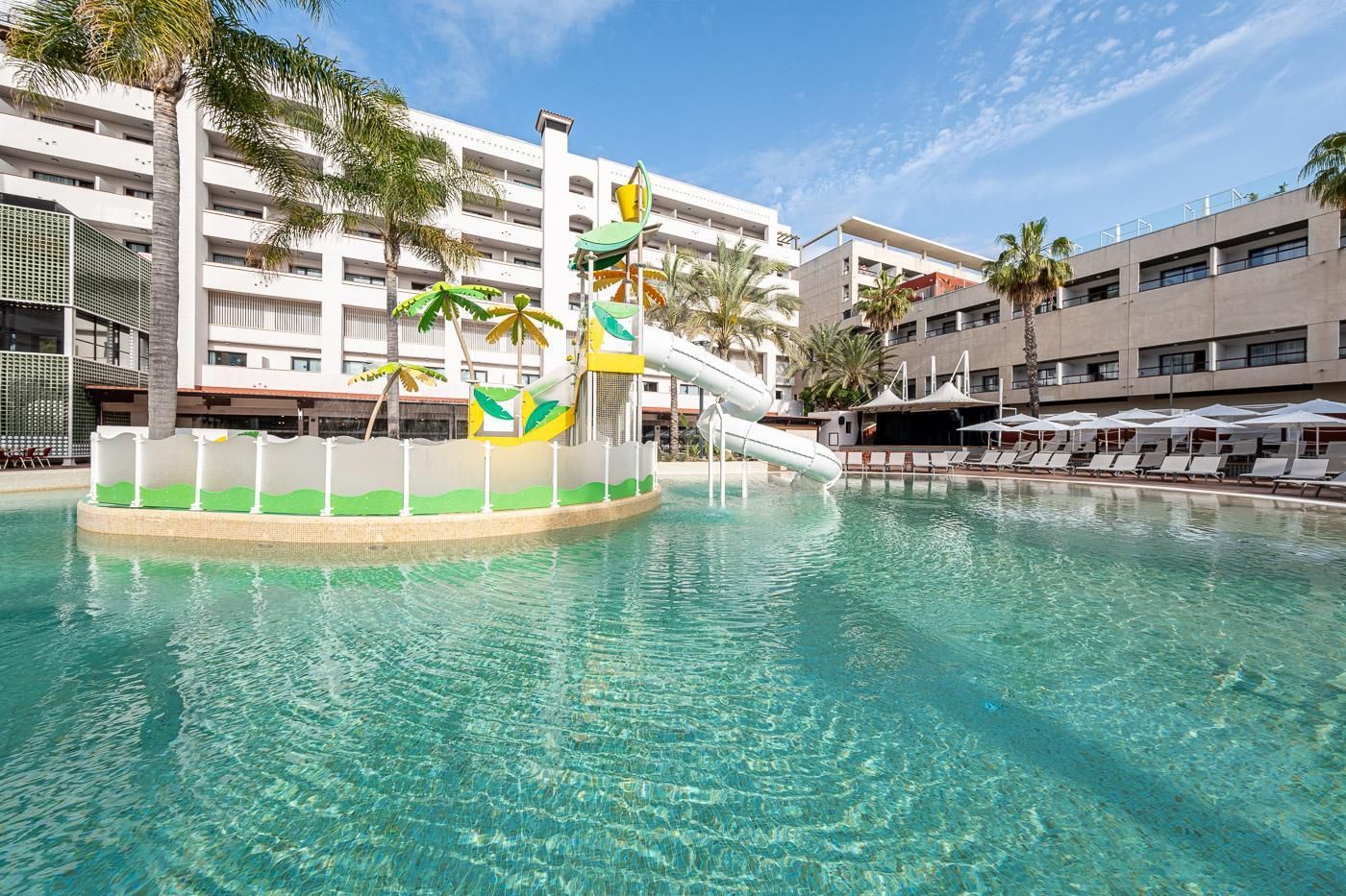 A swimming pool with a slide, palm trees, and a building with multiple balconies in the background.