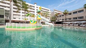 A swimming pool with a slide, palm trees, and a building with multiple balconies in the background.