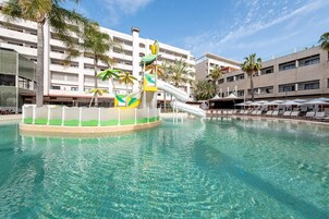 Una piscina con tobogán, palmeras y un edificio con varios balcones al fondo.