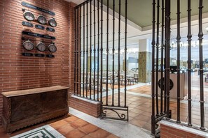 Un vestíbulo con pared de ladrillo, relojes, un banco de madera y vista a un comedor a través de una reja metálica.