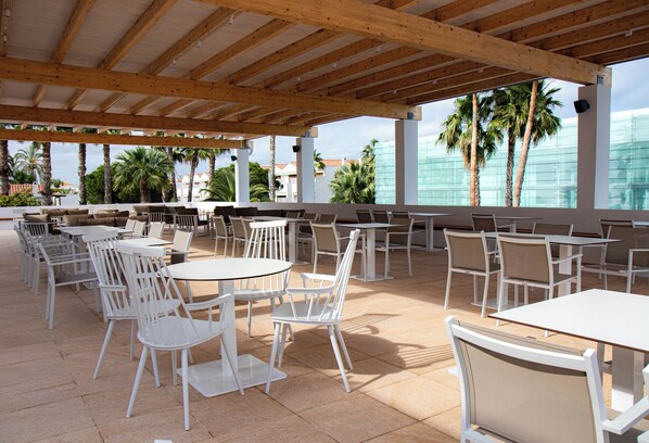 Een terras buiten met witte tafels en stoelen, een houten plafond en palmbomen op de achtergrond.
