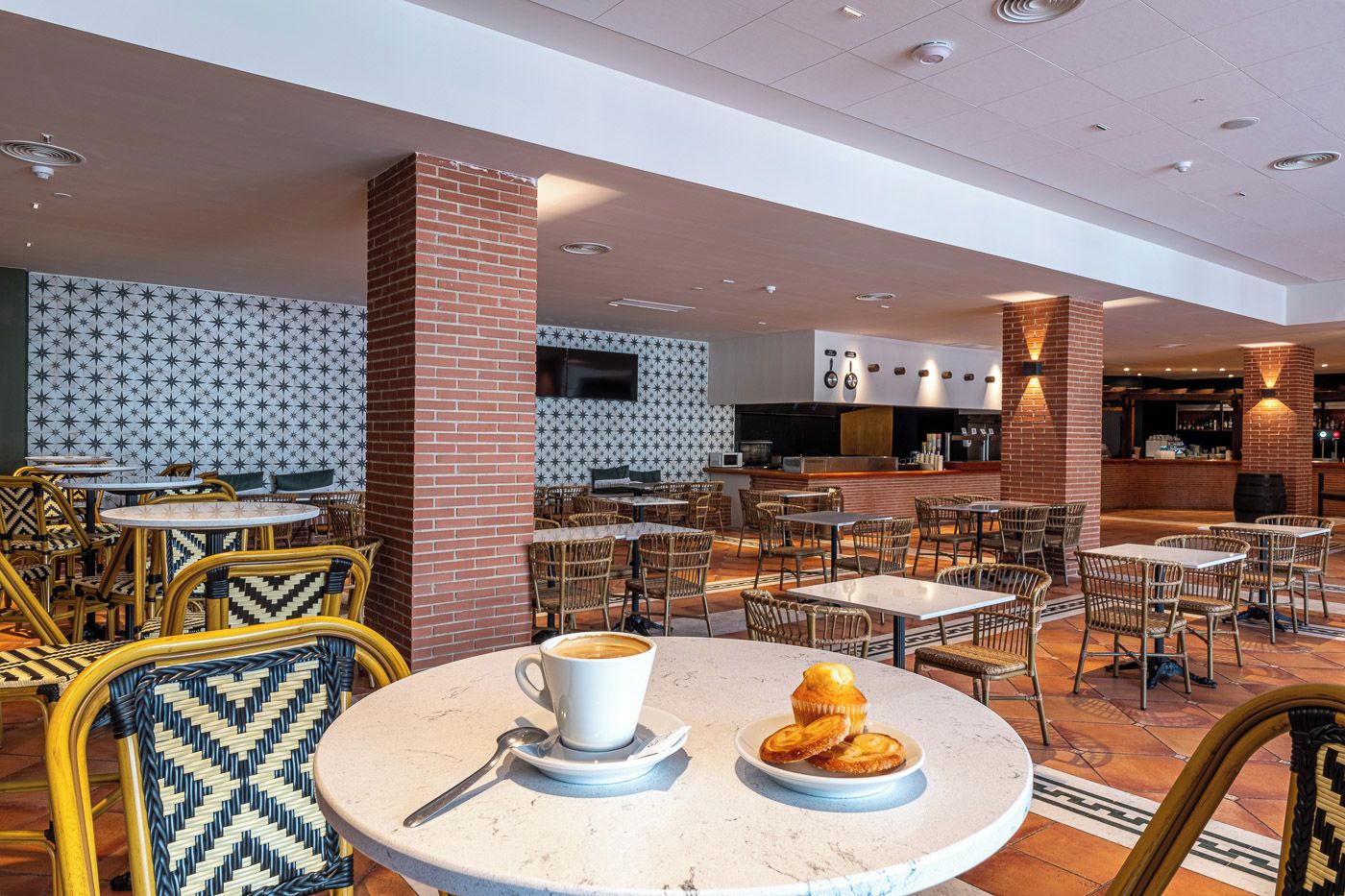 Un café interior amplio con mesas redondas y sillas de mimbre, con un diseño moderno y detalles de ladrillo y paredes con motivos.