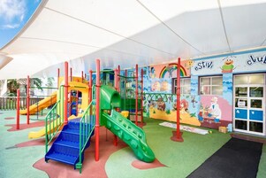 Zona infantil con toboganes y estructuras de escalada de colores, un mural con personajes de caricatura y la entrada de un edificio con un letrero.