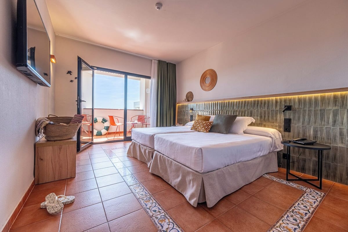A hotel room with two beds, a balcony with chairs, a small table, and a wall-mounted TV.