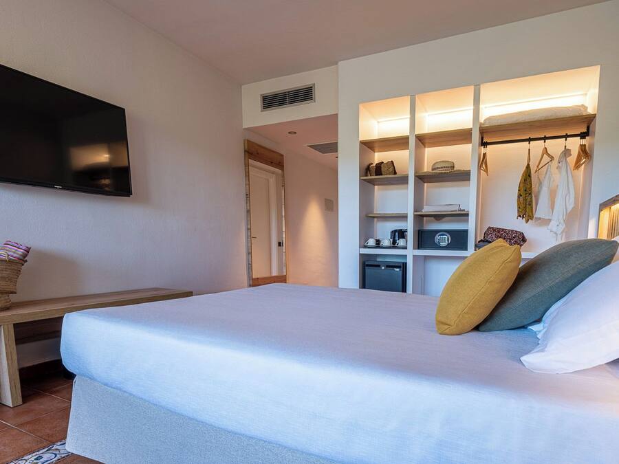 A hotel room with a bed, a TV mounted on the wall, a wooden bench, a small table, a built-in wardrobe, and a kitchenette.