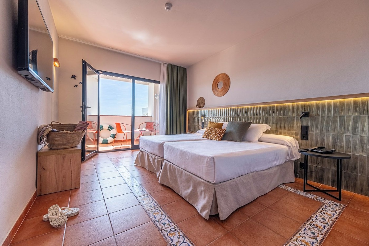 A hotel room with two beds, a balcony with chairs, a large mirror, and a view of the sea.