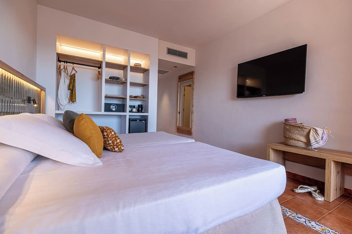 A hotel room with a bed, a TV mounted on the wall, a wardrobe with built-in lighting, and a small table with slippers.