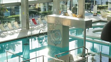 Piscina interna com paredes de vidro transparente, uma coluna decorativa central e áreas de estar iluminadas pelo sol.