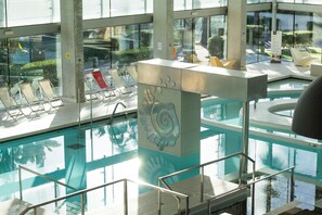 En indendørs swimmingpool med glasvægge, en dekorativ søjle i midten, og solbeskinnede siddeområder.