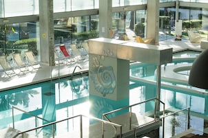 Una piscina interior con paredes de vidrio transparente, una columna decorativa central y zonas de estar iluminadas por el sol.