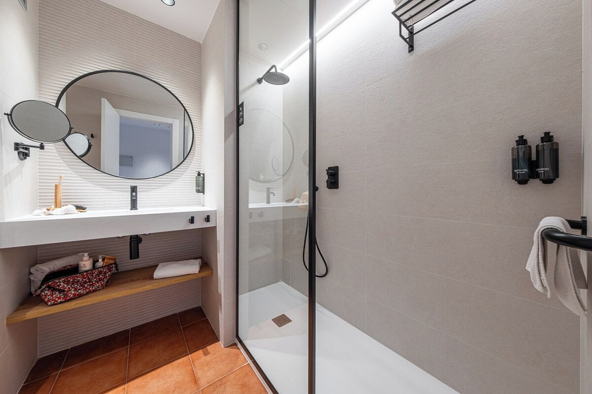 Een moderne badkamer met een douchecabine van glas, een witte wastafel met een ronde spiegel en een handdoekhouder.