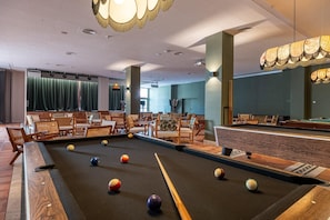 Une table de billard avec des billes, des chaises en bois et une table de jeu dans une pièce aux rideaux verts et dotée d’un luminaire au plafond.