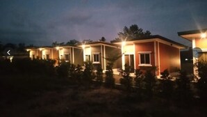 Fileira de edifícios térreos com janelas iluminadas, cercada por pequenas árvores.