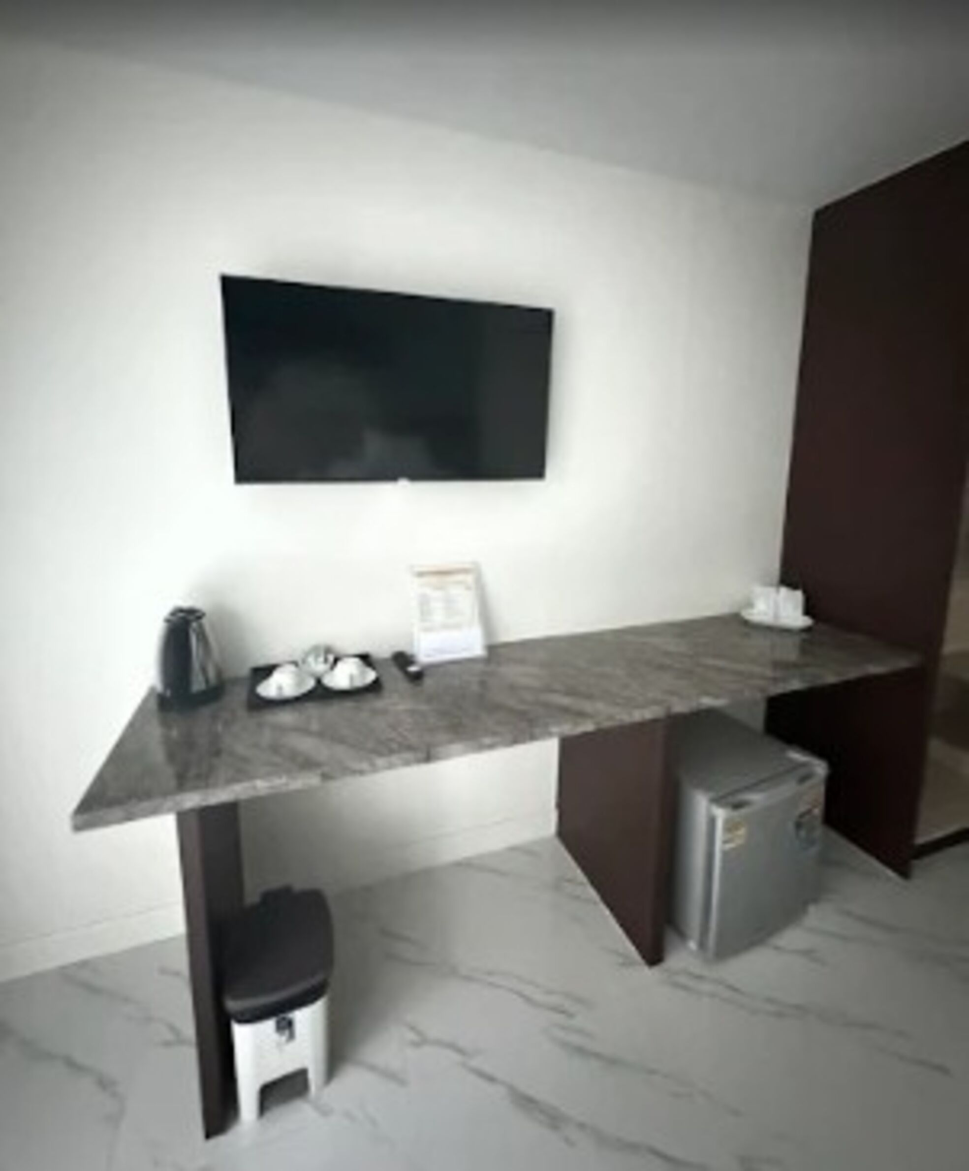 Una habitación moderna con un escritorio de mármol, un televisor de pantalla plana en la pared, un refrigerador pequeño y un cesto de basura.