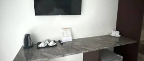 Una habitación moderna con un escritorio de mármol, un televisor de pantalla plana en la pared, un refrigerador pequeño y un cesto de basura.
