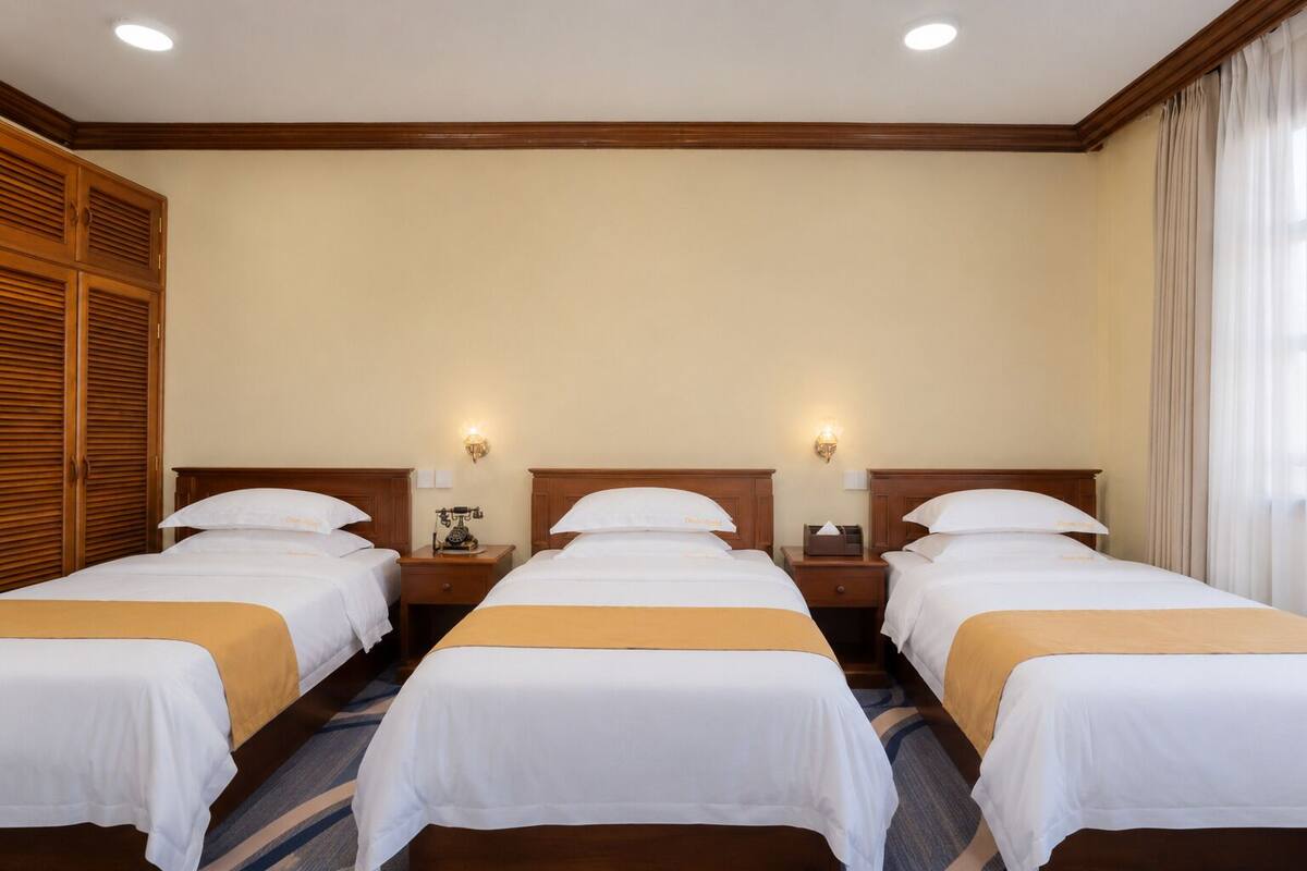 A hotel room with three single beds, wooden headboards, bedside tables with lamps, and a large window with curtains.