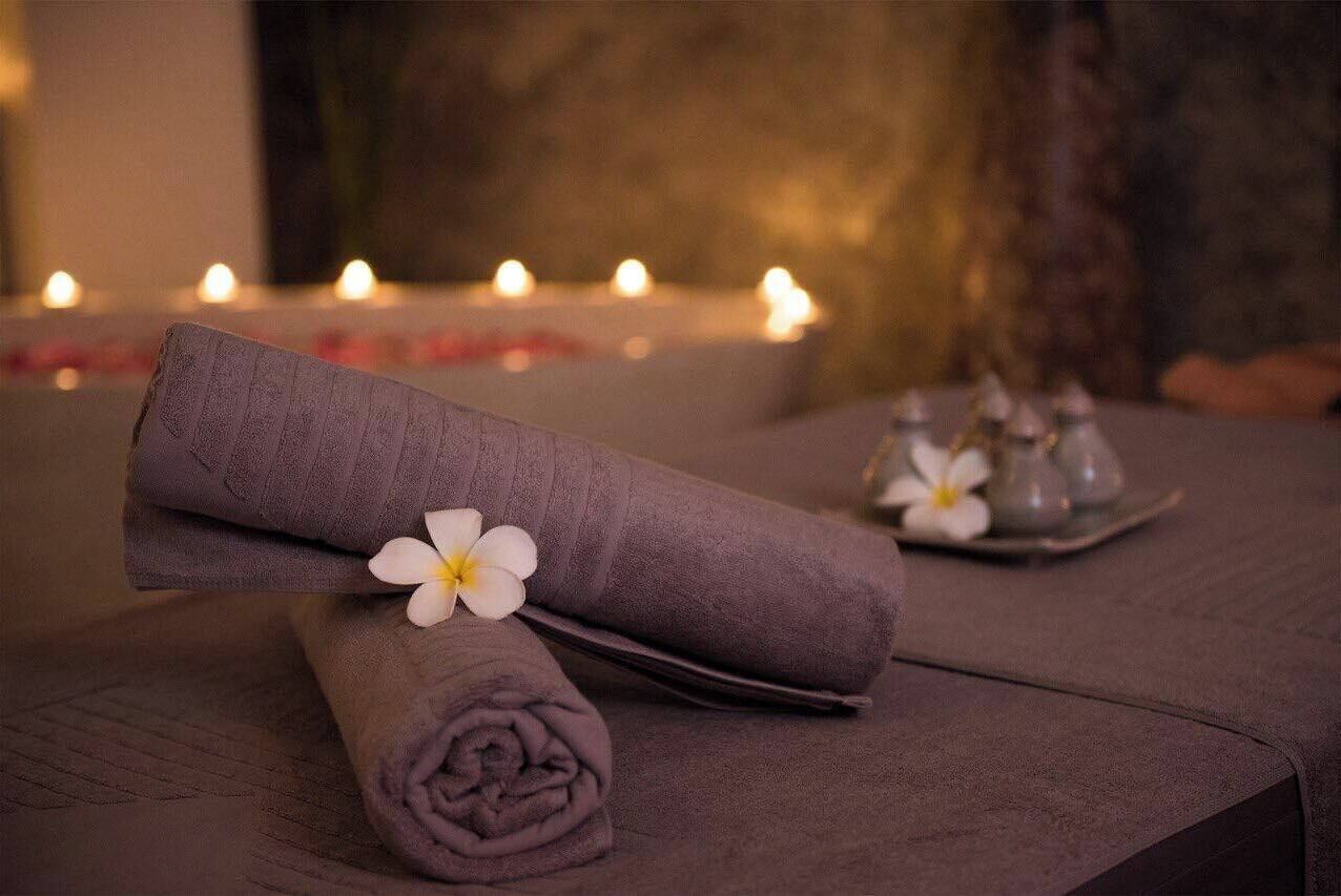 A neatly arranged spa setting with a rolled towel and a flower, candles in the background, and a tray with glasses.