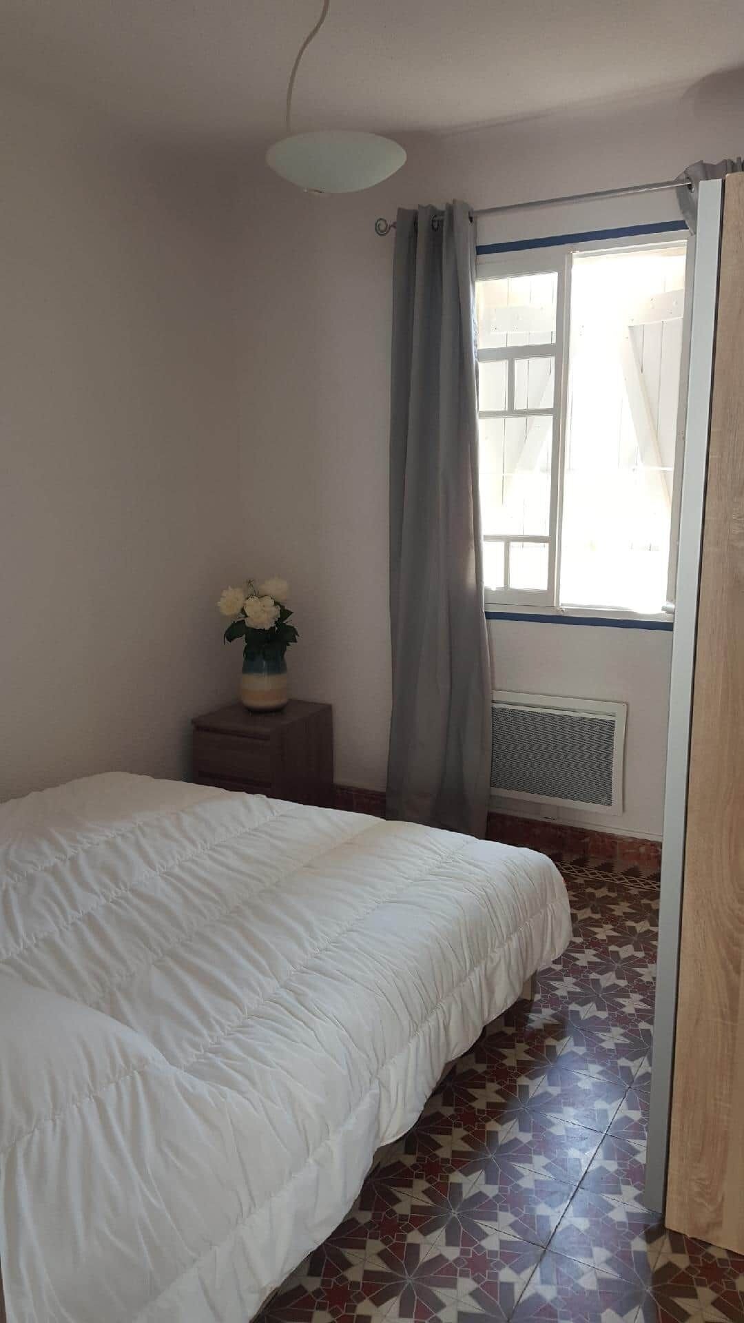 Un dormitorio con cama, una mesita de noche con un jarrón de flores, una ventana con cortinas y un radiador.