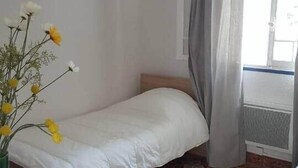 Une petite chambre avec un lit simple, un sol à motifs, une table de chevet ornée d’un vase jaune et une fenêtre avec des rideaux.