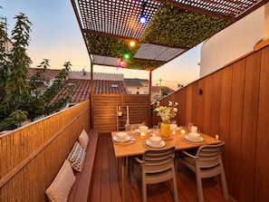 An outdoor dining area with a wooden table set for two, surrounded by chairs, under a pergola with a lattice roof. - Spacious 3-Bedroom Apt • Terrace • Beach 50m (Canet-en-Roussillon)