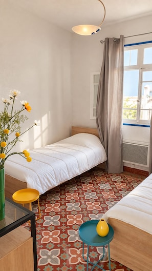 A bedroom with a bed, a small table with a vase, a patterned floor, and a window with curtains. - Spacious 3-Bedroom Apt • Terrace • Beach 50m (Canet-en-Roussillon)