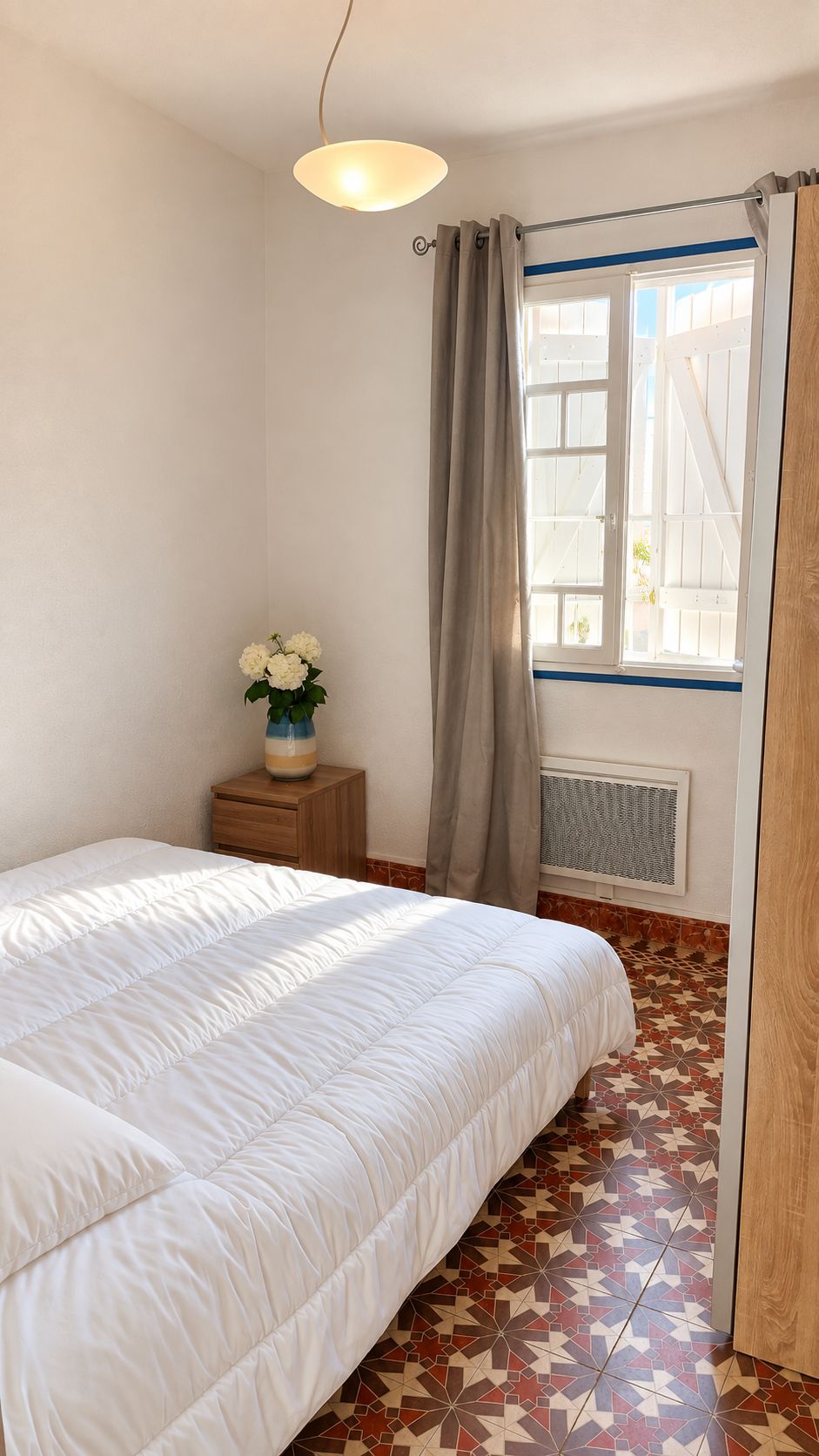 A bedroom with a bed, a bedside table with a vase of flowers, a window with curtains, and a radiator.