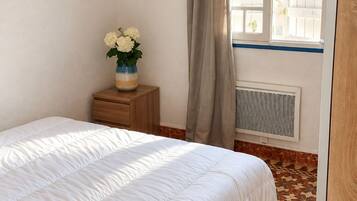 A bedroom with a bed, a bedside table with a vase of flowers, a window with curtains, and a radiator.