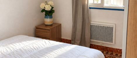 A bedroom with a bed, a bedside table with a vase of flowers, a window with curtains, and a radiator.