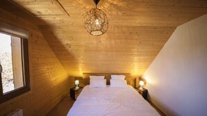 A cozy, well-lit bedroom with a wooden ceiling, a large bed with white linens, two bedside tables with lamps, and a window on the left.