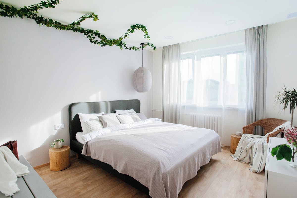 Ein modernes Schlafzimmer mit einem großen Bett, einem Nachttisch, einem kleinen Hocker, einem Korbstuhl, einer Heizung und einem Fenster mit Vorhängen.