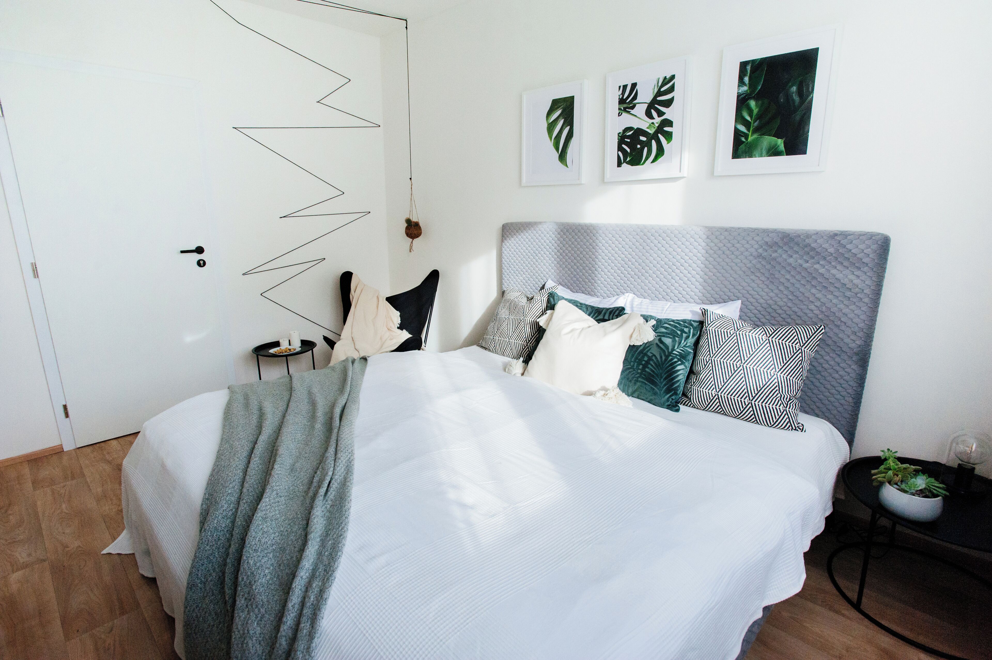 A neatly made bed with a grey headboard, white bedding, and a mix of patterned pillows. There are three framed leaf artworks on the wall, and a small potted plant on a side table.