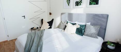 A neatly made bed with a grey headboard, white bedding, and a mix of patterned pillows. There are three framed leaf artworks on the wall, and a small potted plant on a side table.