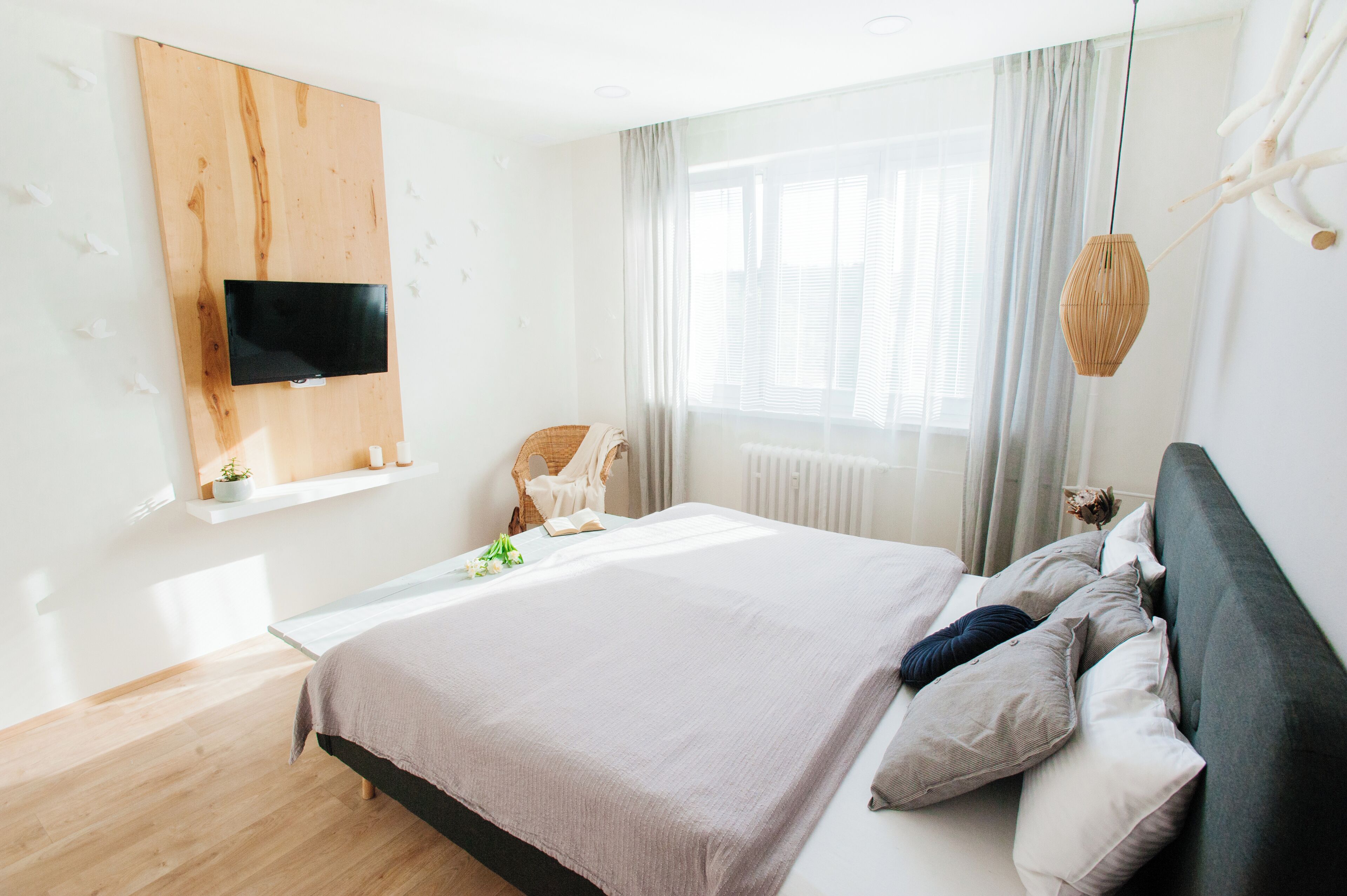 A modern bedroom with a large bed, a mounted TV, a window with curtains, and a bedside table with a lamp.