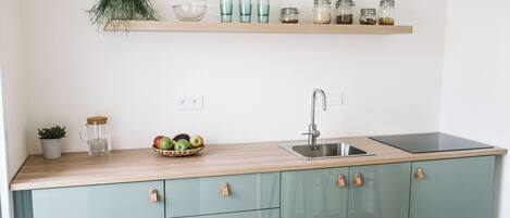 Una cocina con encimera de madera, un fregadero y un armario verde con cajones. Hay botes en un estante sobre la encimera y una planta a la izquierda.