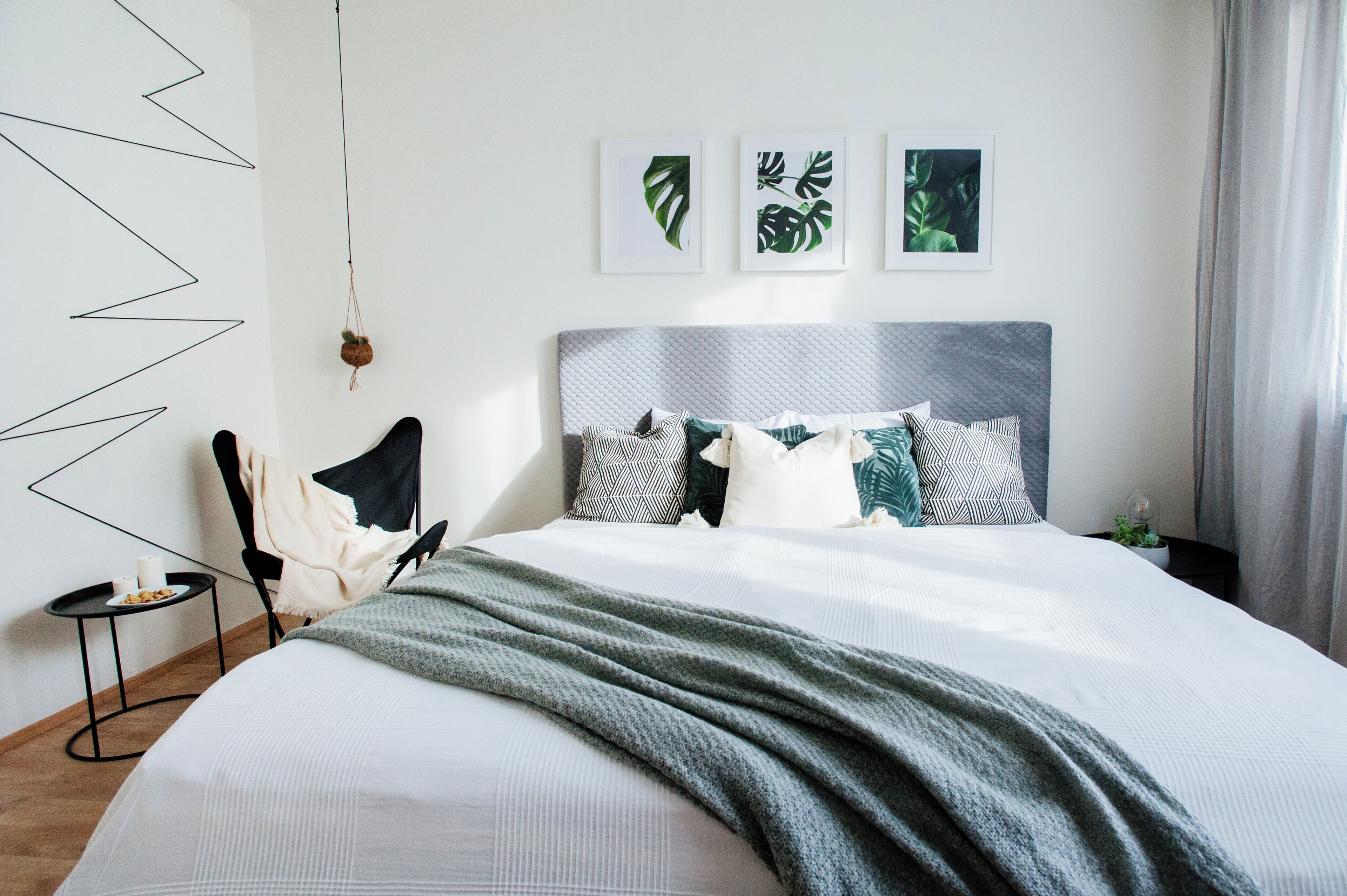 A modern bedroom with a large bed, a side table with a cup and plate, and three framed leaf artworks on the wall.