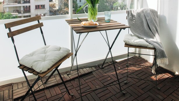 Un balcon doté d’un plancher en bois, d’une chaise blanche avec coussin, d’une petite table ornée d’un vase de fleurs et offrant une vue sur les bâtiments au loin.