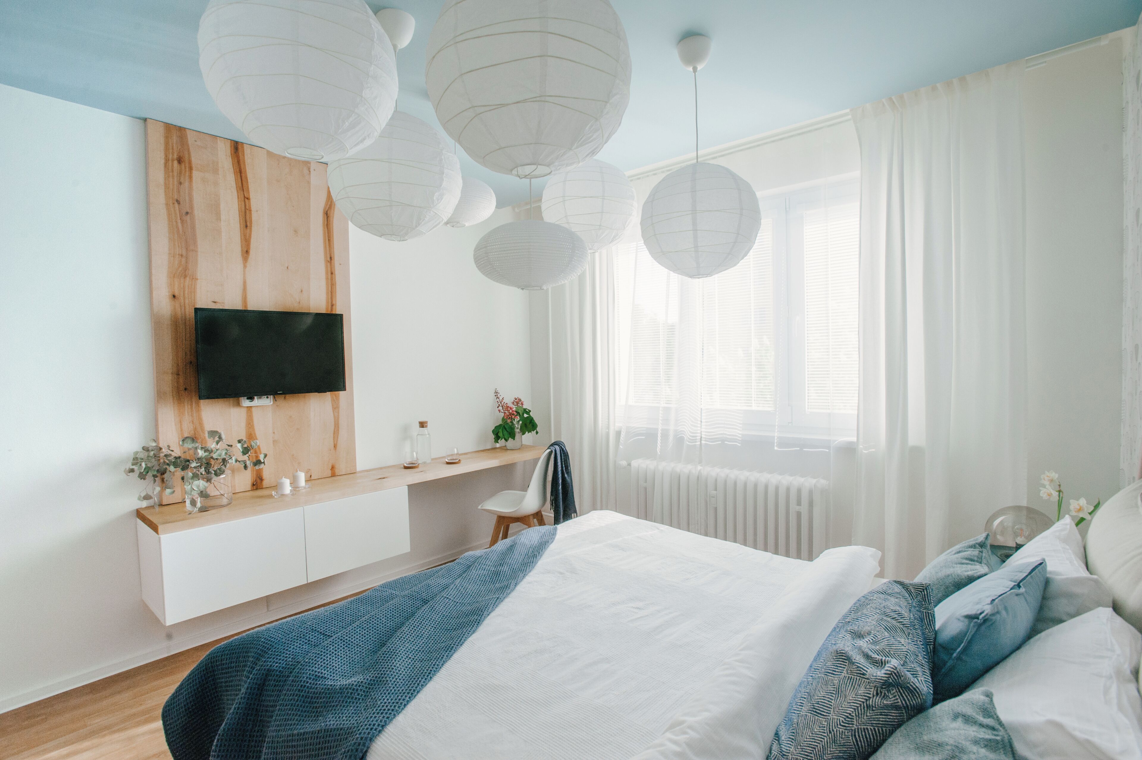 Un dormitorio moderno con cabecera de madera, televisor montado en la pared, mesita de noche con planta y una cama grande con ropa de cama azul y blanca.