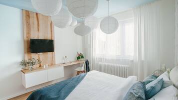 Un dormitorio moderno con cabecera de madera, televisor montado en la pared, mesita de noche con planta y una cama grande con ropa de cama azul y blanca.