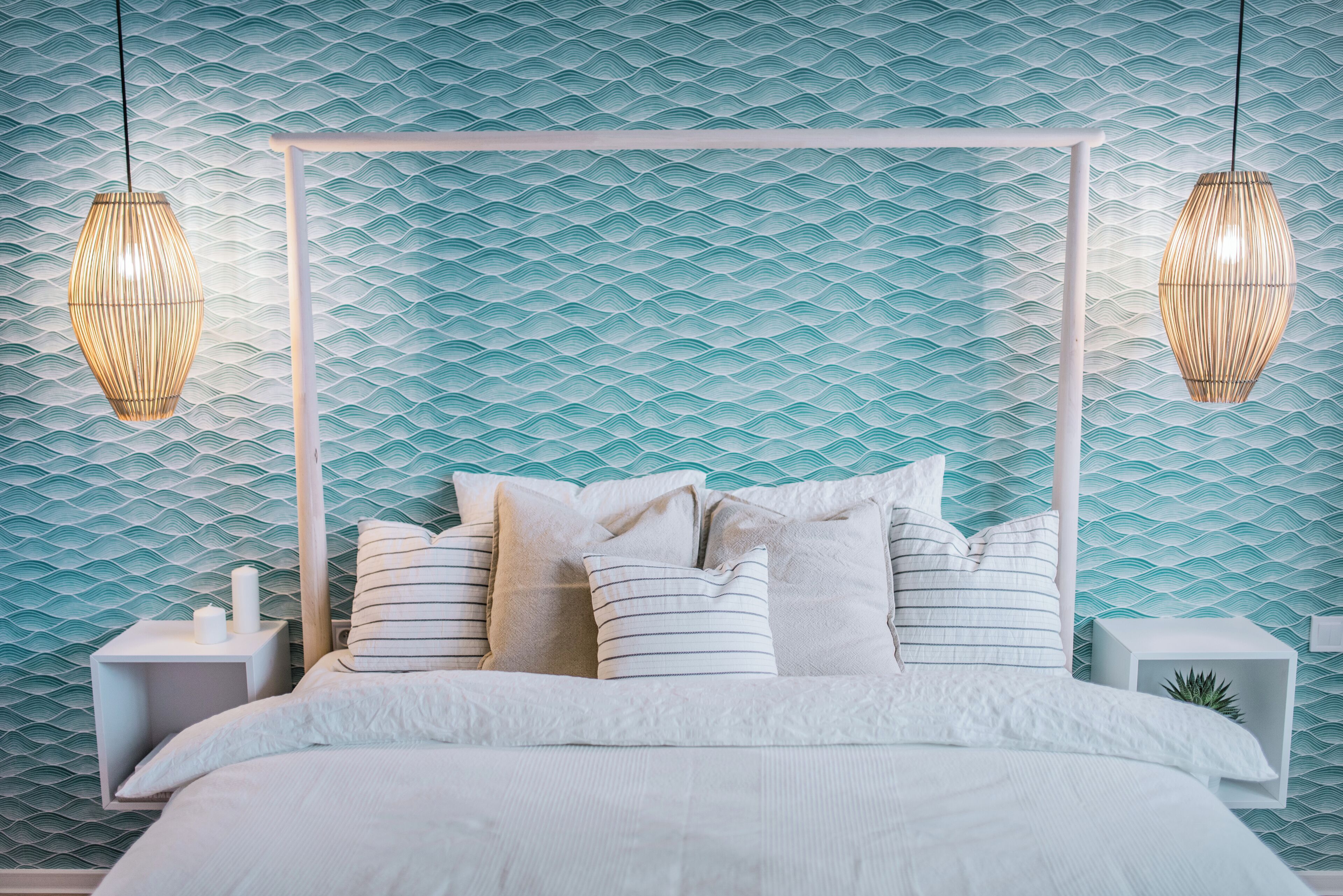 Un lit avec une tête de lit blanche, une literie blanche et des coussins à rayures. Il y a deux tables de chevet avec des lampes blanches, chacune agrémentée d’une petite plante. Le mur présente un motif ondulé.