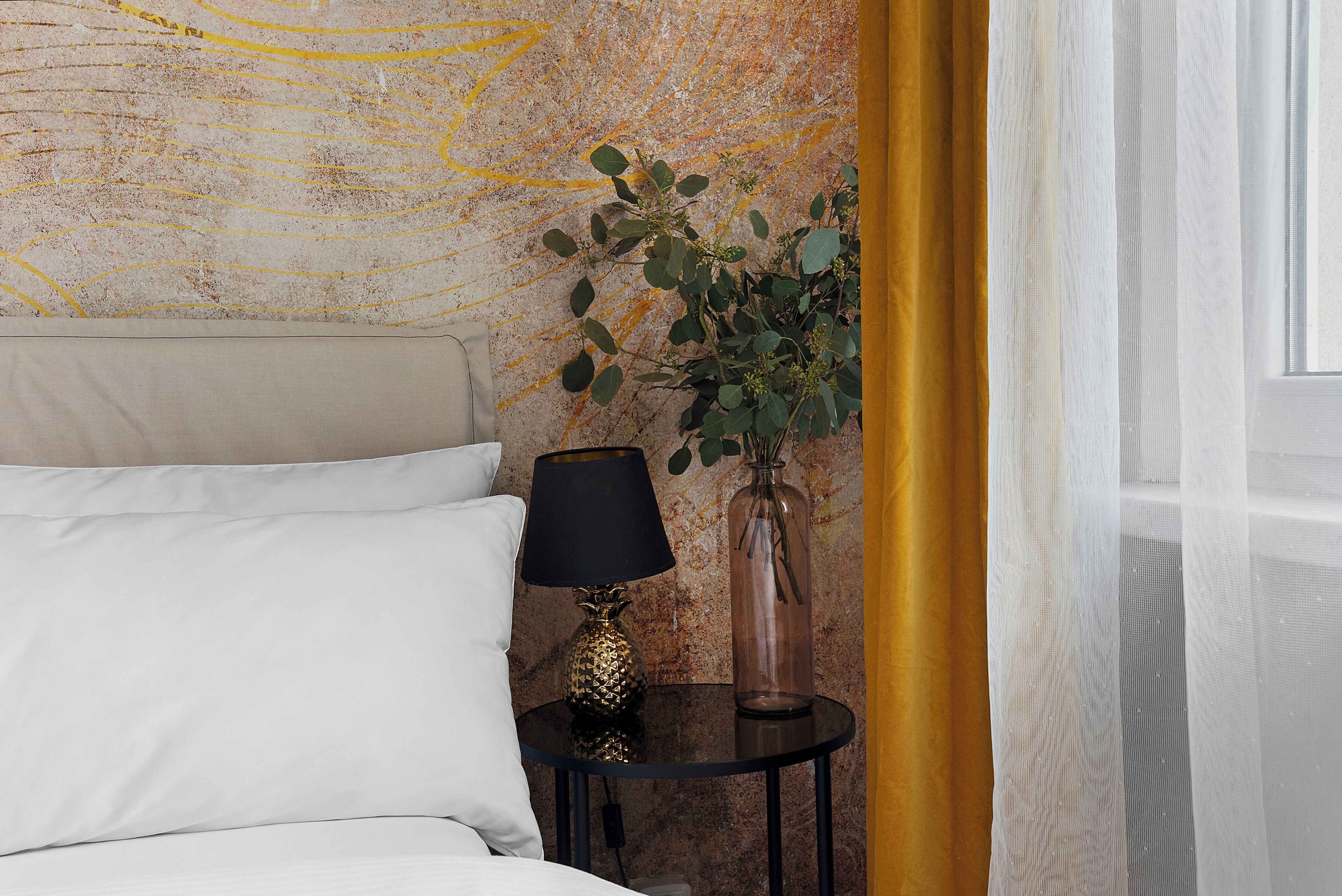 Een bed met witte lakens, een klein rond tafeltje met een lamp, een vaas met groen en een structuurwand.