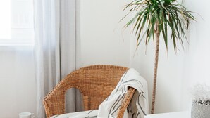 Une chaise en osier avec une couverture blanche, un petit tabouret en bois, une plante en pot et une table blanche dans une pièce au sol en bois clair et aux murs blancs.