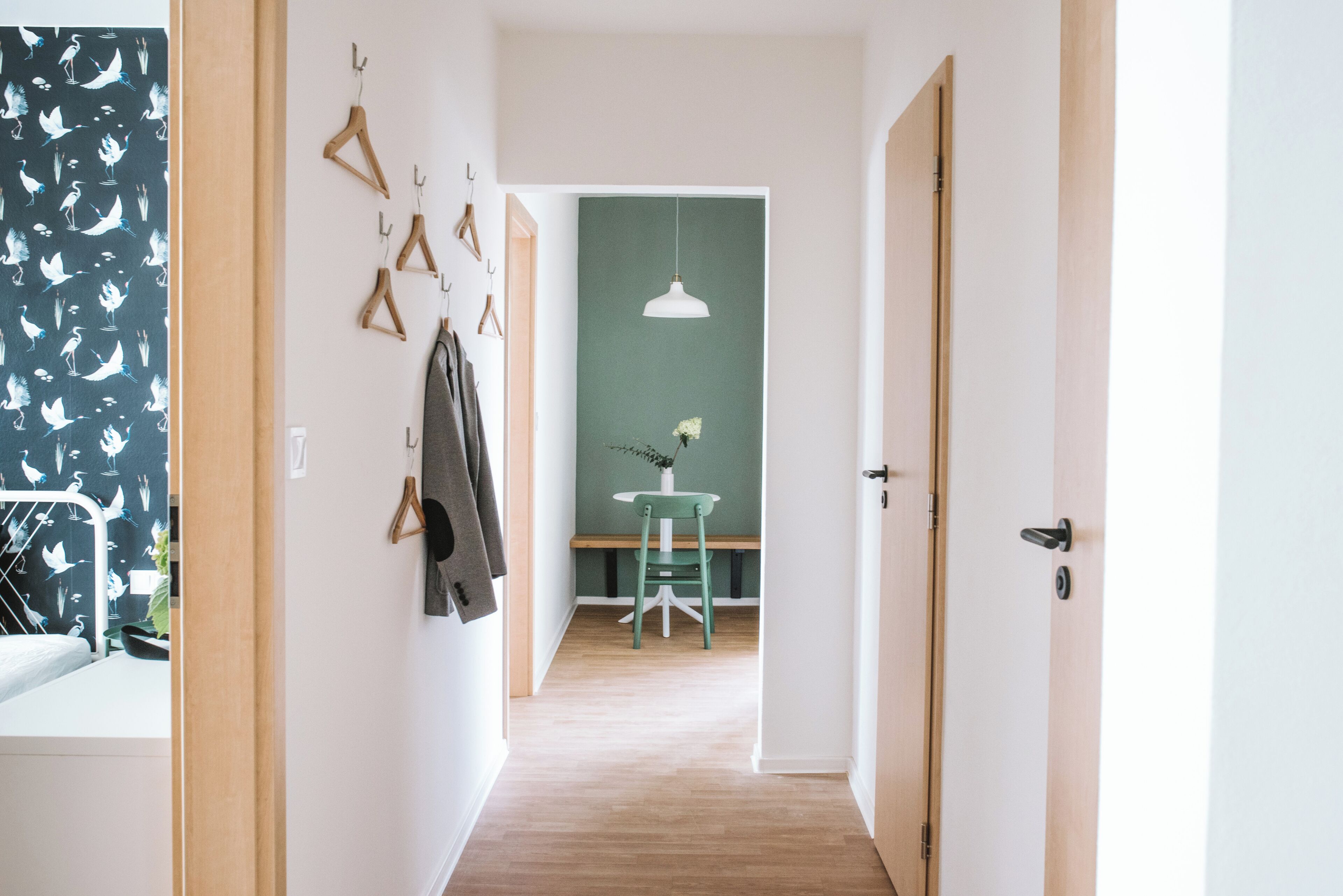 Un couloir avec un plancher en bois, un mur vert, un luminaire suspendu blanc et une table avec une chaise.
