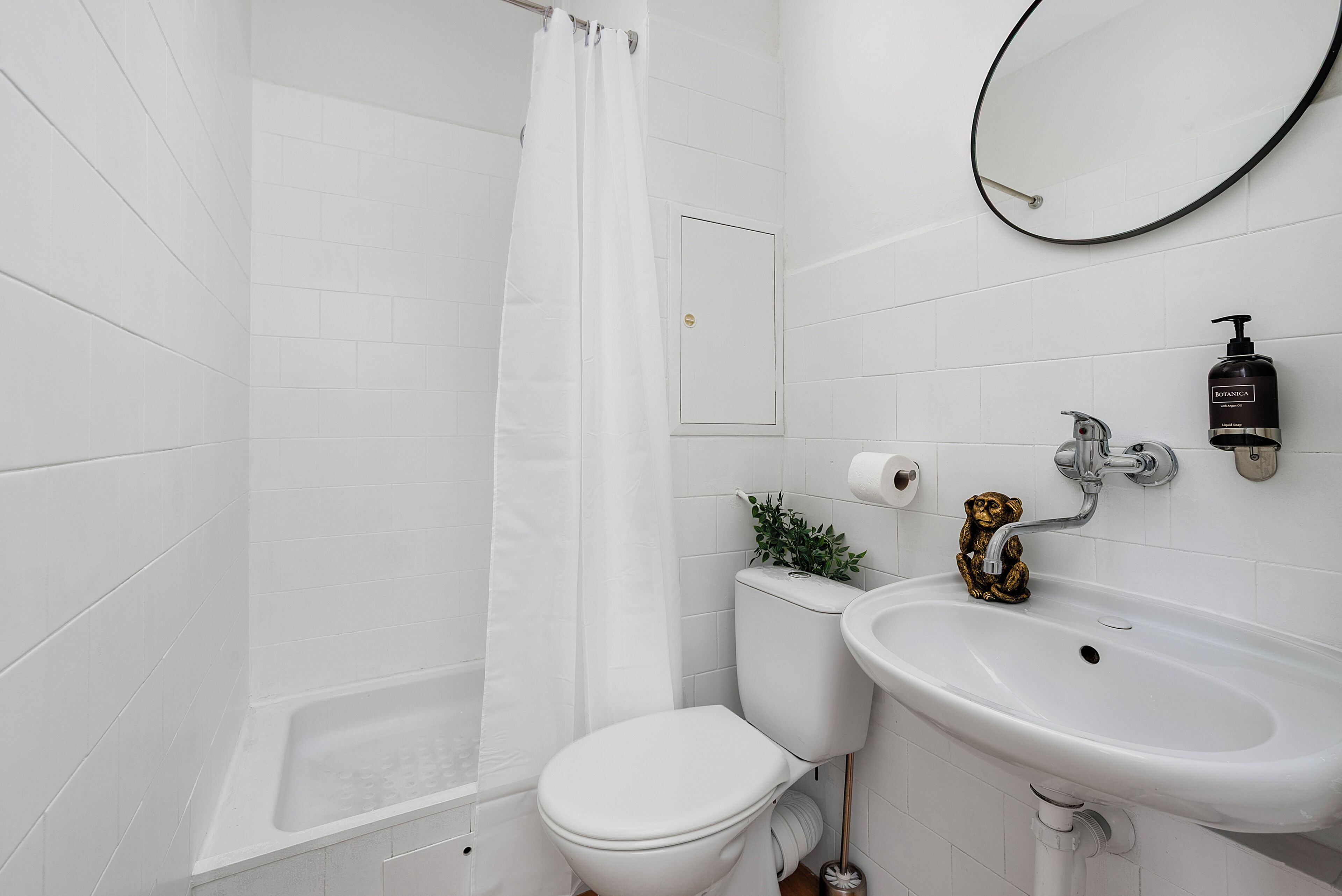 Et badeværelse med hvide fliser, badekar, toilet, vask og et spejl, samt en lille plante.