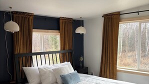 A bedroom with a large bed, two bedside tables, a nightstand, a hanging lamp, and a window with curtains. - Barrel Sauna + Open Concept + Mountain Getaway = Relaxing Stay (Wasilla)