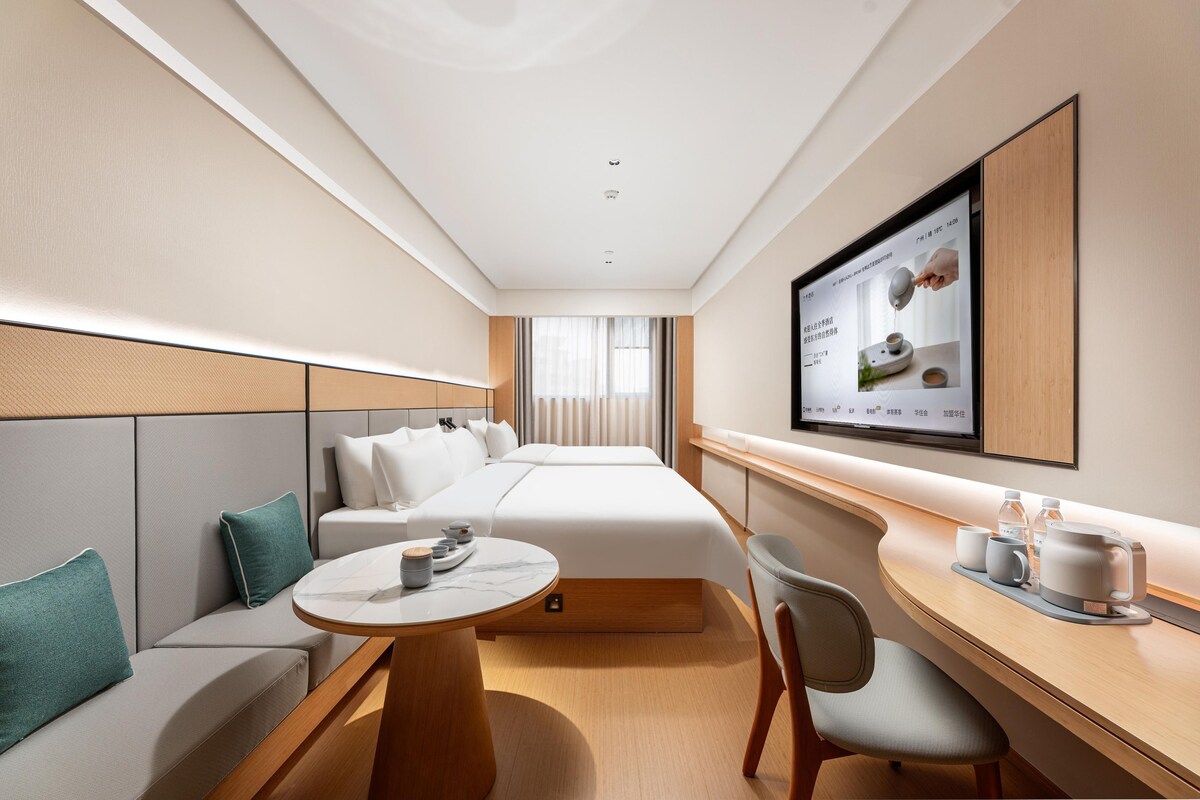 A hotel room with a bed, a seating area with a table, a TV mounted on the wall, and a window with curtains.