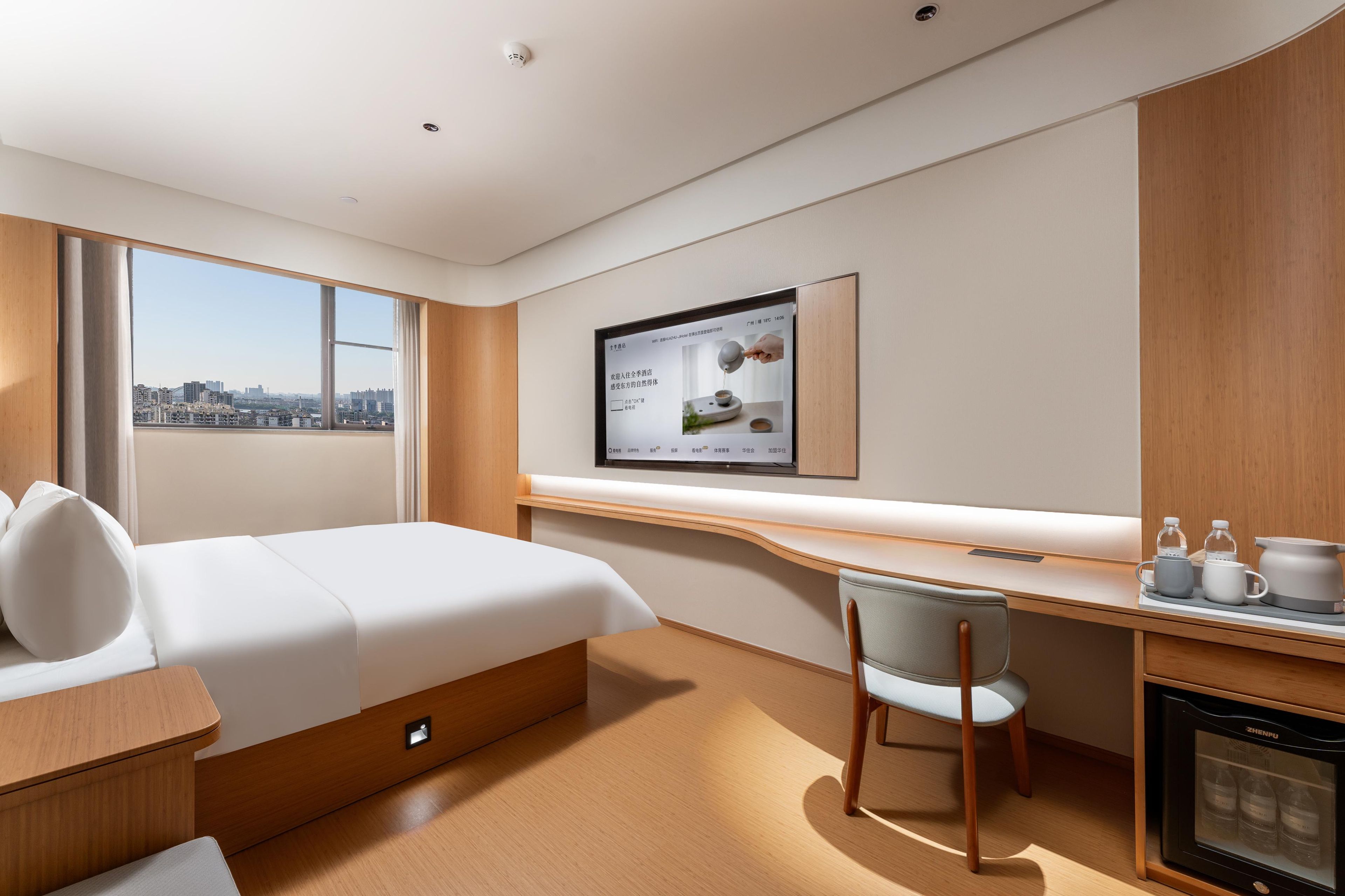 Habitación de hotel con cama, escritorio, silla, un refrigerador pequeño y ventana con vista a la ciudad.