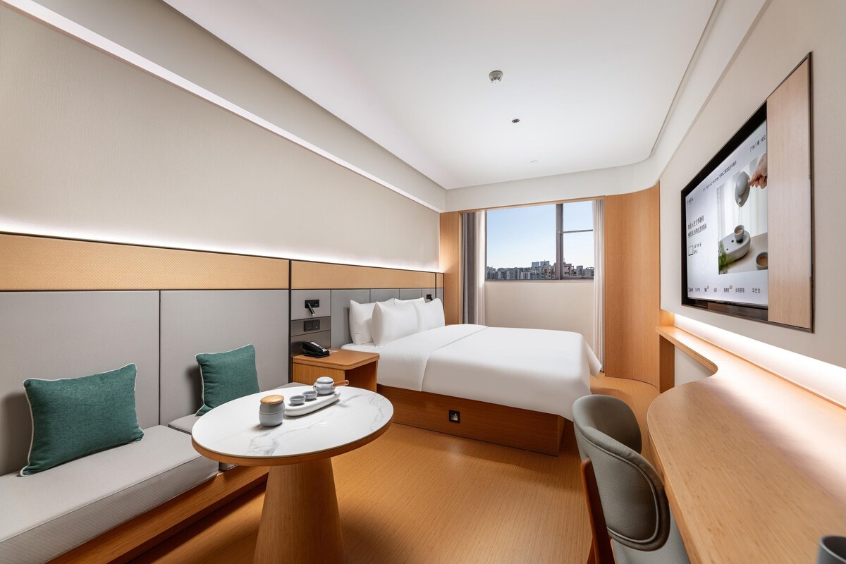 A hotel room with a bed, a desk, a chair, a TV, and a view of the city.