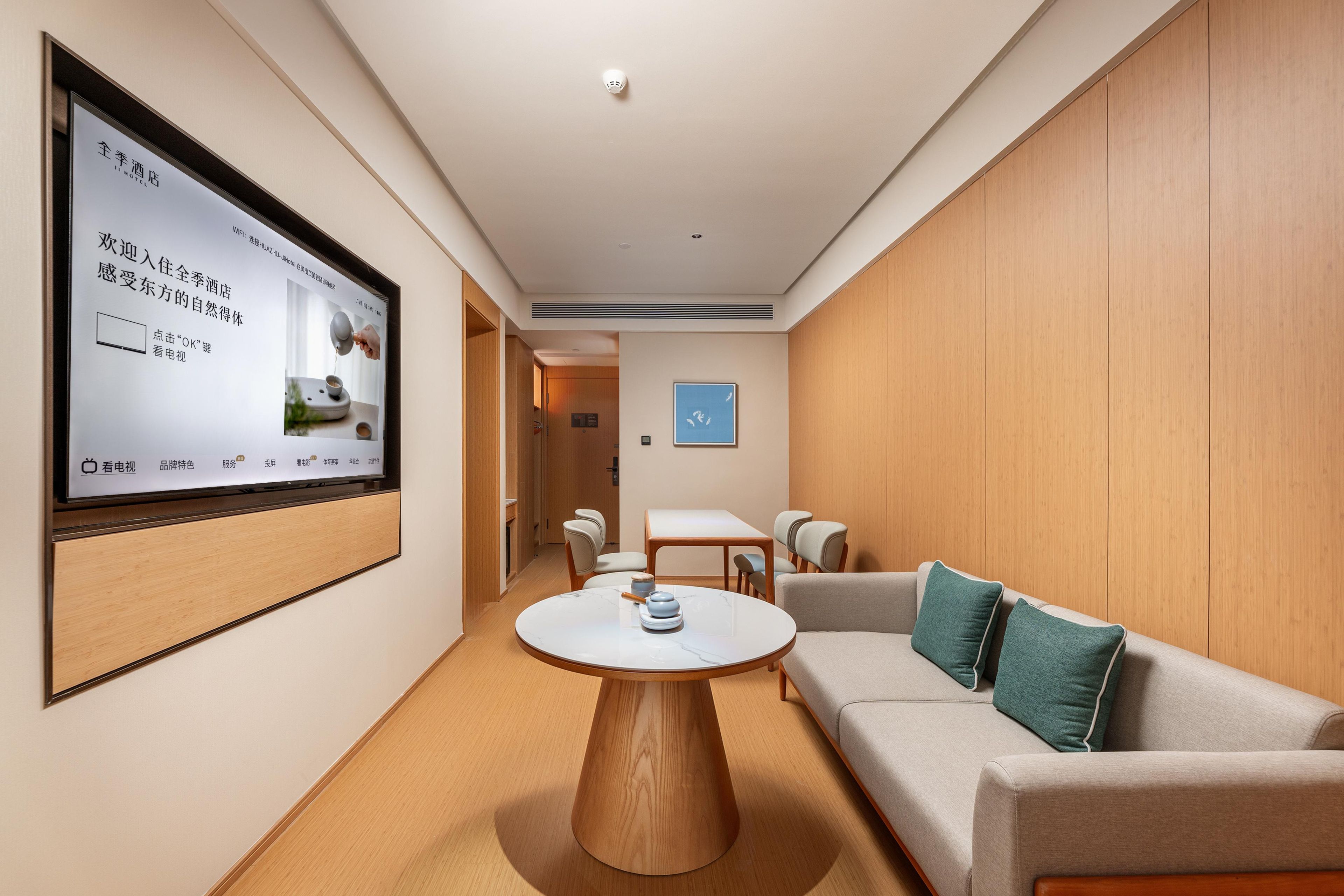 Una habitación de hotel moderna con una pantalla grande mostrando información, una mesa de centro redonda de madera, un sofá con cojines y un diseño minimalista.