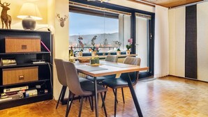 Ein Essbereich mit Tisch, Stühlen, einer Kandelaber, einem Bücherregal und Blick durch ein großes Fenster.