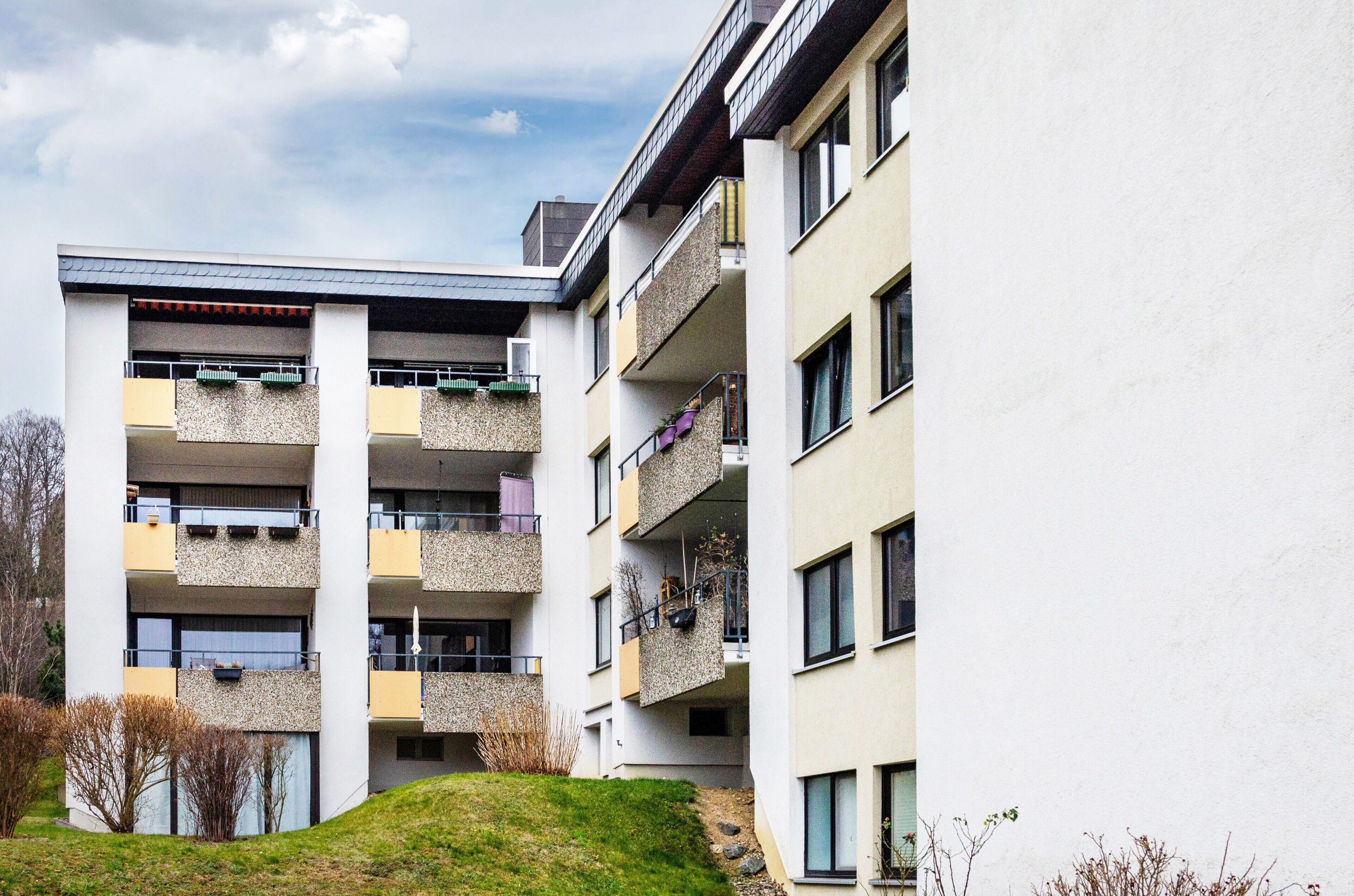 Un edificio residencial de varios pisos con balcones, techo plano y un área verde en la parte delantera.